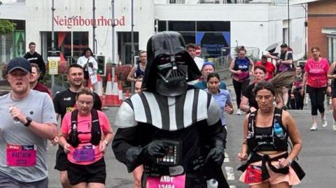 A person dressed as Darth Vader from Star Wars is running in front of other men and women, who are also competing but wearing shorts and T-shirts.