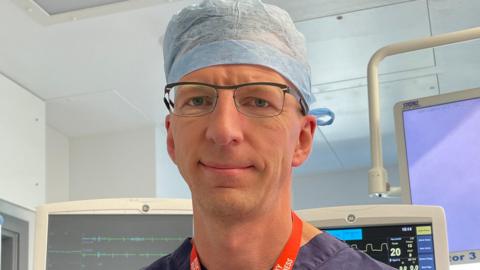 Leo Krivskiy looks at the camera, he is in a surgery theatre room with screens showing medical date behind him and wears blue scrubs. He has a red lanyard around his neck, dark blue scrubs and a pale blue hat on, with thin black wire framed glasses.