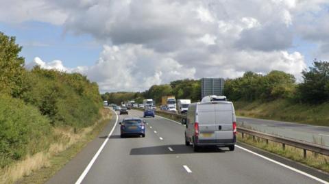 The A14, showing two dual carriageways, with vehicles on the road. A central reservation, a traffic sign in the distance, bushes and grass either side, a large sky, with grey clouds in the sky. 