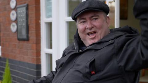 Tony Swift outside a block of flats. He is wearing a black cap and dark coat