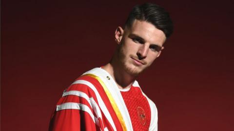 Footballer Declan Rice stood looking at the camera with his dark hair slicked back over his head. He is wearing Arsenal's home kit which is mainly red with some white and has a red and white Arsenal flag draped over his right shoulder. The background is just a red-black colour.