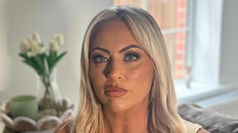 A woman with long blonde hair and blue eyes is looking directly into the camera, unsmiling. She is wearing a light-coloured sleeveless v-neck top. The background is out of focus but she is sitting on a grey sofa with light green cushions, behind her is a table with white flowers on it and a window. 