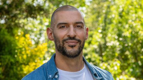 A man wearing a white t-shirt and a blue denim jacket smiling, with leafy green trees visible behind