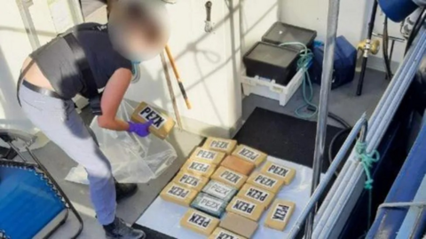 A person with a blurred face picks up brown packages about the size of a brick, and puts them into a clear plastic bag, while standing on the deck of a boat