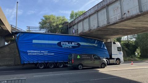 A white cab and a blue trailer on a road. The articulated lorry's trailer is stuck under a grey bridge.