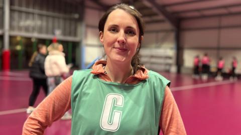 Sammy smiles at the camera. She's standing in front of a netball court and some players are getting ready behind her. She's waiting a pink, zip up fleece with a light green bib with the letter C on it. Her dark hair is tied back in a ponytail.