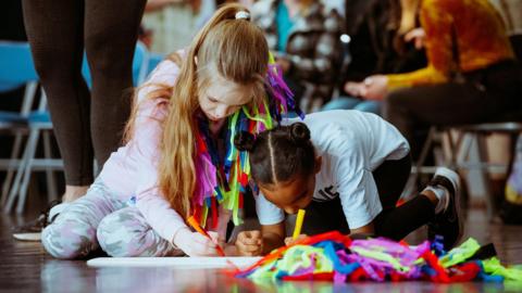 Children draw on paper with crayons. The paper is resting on a wooden floor in a hall, and there are chairs visible in the background