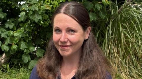 A young woman with long dark hair over both her shoulders wearing a dark blue top with flowers on it, standing in front of a hedge in a garden.