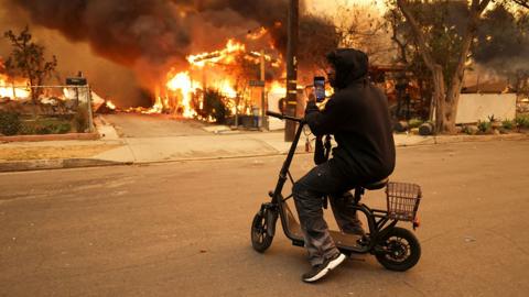 A person uses a cellphone to take a picture of a burning building while sitting on a stationary scooter with their hood up at the Eaton Fire in Altadena on Wednesday.