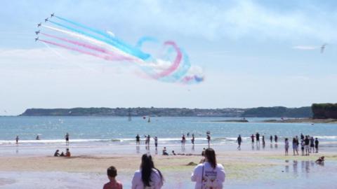Crowds lining the beach as five planes fly next to each other in the sky with red white and blue smoke coming out behind.