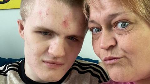 A young man pictured beside his mother, this is a close up picture of their faces. The teenagers t-shirt has three black stripes on his shoulders with a grey background, his mothers top is pink.