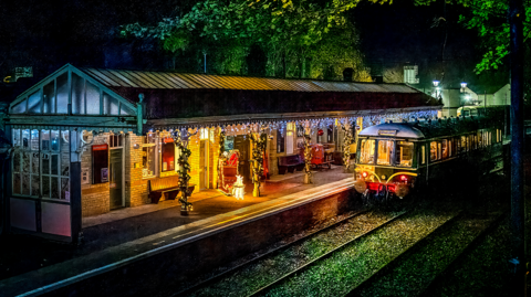 The green heritage train is pulled up to the station, which is adorned in Christmas decorations including lights and Santa's sleigh.