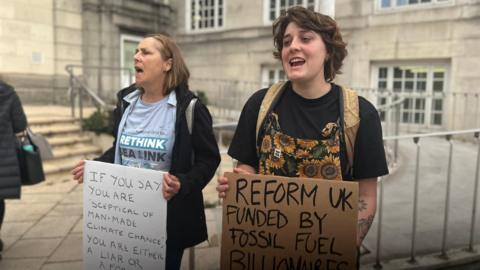 Two women stand outside Kent County Council's offices in Maidstone holding placards. One says "if you say you are sceptical of man-made climate change you are either a  liar or a fool and certainly a coward", the other says "Reform UK funded by fossil fuel billionaires." 