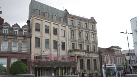 A building of at least four storeys is in the centre of the photo. Smaller buildings are next to it, either side, on the image.