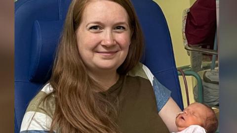 Kate wearing a green t-shirt- holding her baby who is sleeping and has a tube under her nose