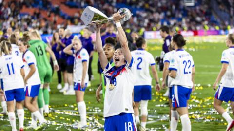 Chloe Kelly celebrates with the Euro 2025 trophy on the pitch with her team in the background.