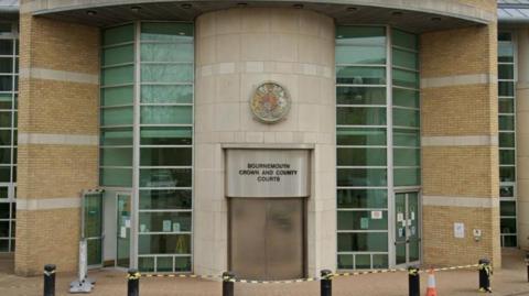 The front of Bournemouth Crown Court. A sign above a large double door at the front of the building reads "Bournemouth Crown and County Courts". Either side of the door are tall strips of windows. In front of the building there is a set of bollards.