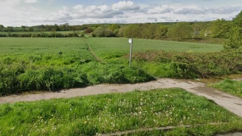 A field covered in grass, with a footpath and the front of the image. The field has a public right of way cutting across it, with trees and hedges surrounding the land. Three sets of floodlights can be seen in the distance. 