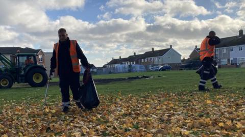 Two workers in orange hi-vis jackets are litter picking among leaves on a Hartlepool field. A green tractor is in the background putting rubbish in a large trailer.