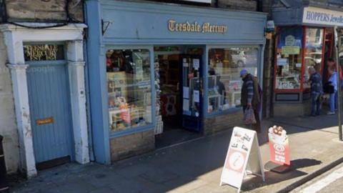 A couple stand outside the office and shop of the Teesdale Mercury and look through the window. There are two large windows either side of the door displaying various goods. The shop front and a door to the side are painted in a blue/grey colour. 