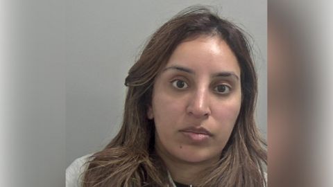 A police mugshot of a woman with long brown hair and a grey top. She is standing in front of a grey wall.