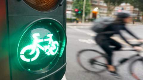 A green traffic light signal illuminates a bicycle on a traffic light. In the blurred backshot is a cyclist on a road.