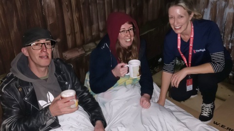 A Threshold member handing out warm drinks and talking to people in Swindon. The threshold worker is wearing a charity top and crouching down, smiling.
A woman and a man are under a duvet holding hot drinks and smiling. The woman is wearing a red hoody and glasses and the man is wearing a dark cap and glasses.
