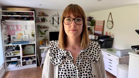 Natalie Ramsell-Lee. She has short brown hair, a fringe and wears glasses. She is also wearing a black and white polkadot blouse. She is sat her desk, with one of her designs on the screen, and is smiling at the camera.