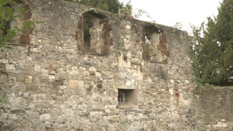 The surviving wall of the 12th Century Bishop's Palace in Halling which is a pale stone wall with rounded windows at the top and a small square window near the bottom.