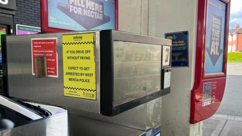 A yellow sign is stuck to the side of a screen advising motorists of the cost of petrol. The sign, in black writing, warns drivers they will be arrested if they fail to pay for their fuel