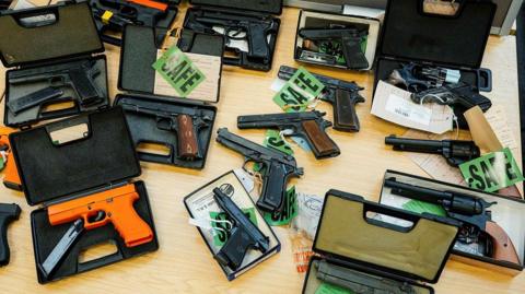 A selection of handguns displayed in boxes on a wooden table. Some are marked with green cards with the word 'safe'
