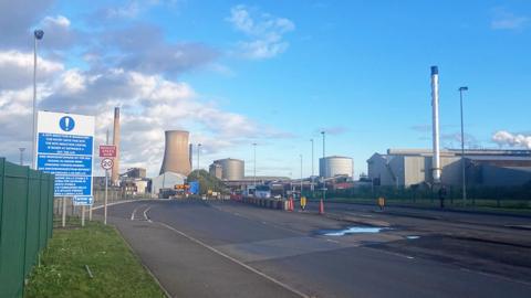 One of the entrance gates at British Steel Scunthorpe with warning signs and buildings, and a large chimney in the background.