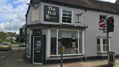 The Nail Room salon is on the corner of a street, the building is painted grey, with a large white framed window and a black painted front door which has an 'open' sign on it. There is a black sign with 'Welcome' written in gold lettering abover the door and a larger square sign which says 'The Nail Room' on the top floor of the two-storey building in gold lettering, with a phone number underneath it. The sign is next to another window. There is a traffic light to the right of the image and on the left is a road with some greenery and housing in the background and a mustard yellow car and a mini behind it on driving on the road