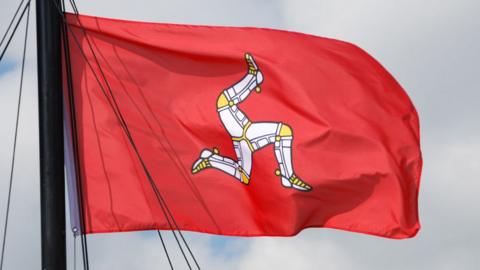 The Isle of Man flag, coloured red with the triskelion in the middle, flies in the wind. The triskelion is an image of three legs in armour, fitted with golden spurs.