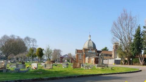 Henley Road Cemetery with road curing around an area of graves with numerous headstones on a grassed area and a building to the right.