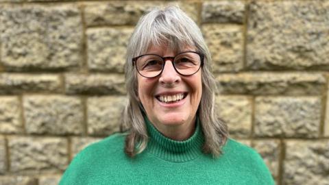 Juliet Layton wearing a green jumper and smiling at the camera in front of a brick wall