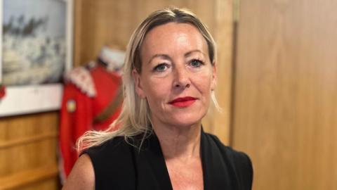 Claire Lilly dressed in a black sleeveless dress stands in front of an old military uniform at Newcastle Territorial Army barracks. She has blonde, shoulder-length hair and bright red lipstick.