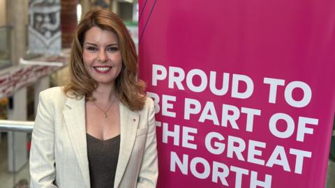 North East mayor Kim McGuinness smiling next to a bright pink banner reading Proud to be part of the Great North at the launch of the Great North in Leeds. She is wearing a white suit jacket and a brown top. Her blonde hair reaches her shoulders and is curled at the ends. Her eyes are brown.