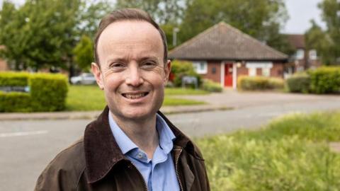 Richard Rout smiles at the camera. He has short brown hair and wears a brown jacket with a blue shirt underneath. He is standing in front of a patch of grass with a bungalow behind him. 