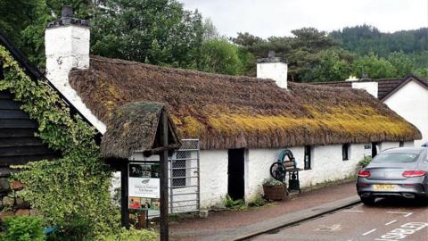 The Glencoe Folk Museum which is a single-storey white building with a thatched roof with a car parked in front of it and trees behind it