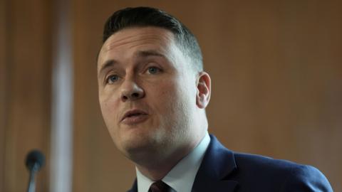 A head and shoulders shot of Wes Streeting mid-speech. He is wearing a navy suit, a white shirt and a navy and red striped tie. 