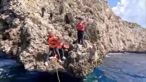 A group of people wearing life-jackets stand on a rocky outcrop with one holding a buoyancy ring