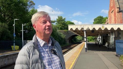 Bruce Duncan is standing on a train platform smiling at the camera. He is wearing a checked shirt and a puffer coat and has pale grey hair. There is no-one else on the platform on this sunny day.