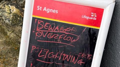 A St Agnes RNLI sign leans against a wall. In red chalk on a black background is written sewage overflow, following by Lightning.  