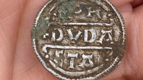 A close-up of one of the coins in the palm of someone's hand. It is silver with writing on and has mud on it.