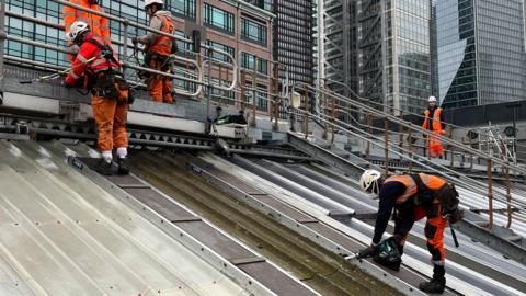A man in hiviz and a helmet is tied to a rope and on a roof. He is cutting a section of green sheeting from the roof.