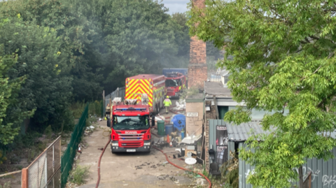 The image shows about three fire engines at the side of a factory site with hoses coming from them amid debris. There is a chimney on the side of the factory with corrugated buildings/rooves in the foreground with a tree also in front of them.