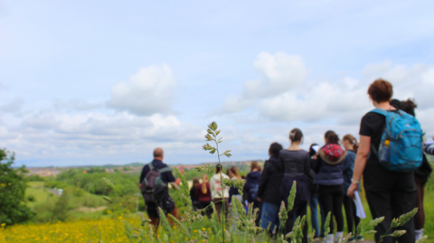 A group of young people walking at Rainton Meadows Nature Reserve. They are walking down a path which is surrounded by green space. Some have backpacks on their backs. The sky is blue with white clouds.