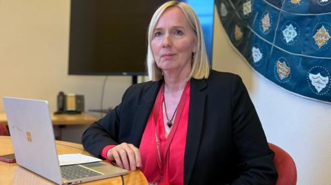 Catherine sits by a wooden table in a red, velvet chair. She's wearing a black jacket with a red blouse. There's a laptop on the table in front of her with a piece of paper on and her phone is next to it. She has short blonde hair and blue eyes. There's a large flat screen TV behind her.