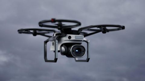 A grey and black drone hovering in the sky. It has four 'wings' as part of it.
